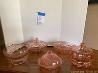 Five pieces of pink depression glass arranged on a wooden surface, including covered dishes with knob handles and bowls with decorative patterns.