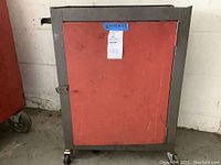 Front view of the rolling metal cabinet, showing red door with hasp lock and exterior dimensions tag