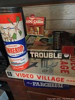 Photo showing Tinkertoy tin container, miniature Log Cabin syrup container, Trouble board game, and other games stacked