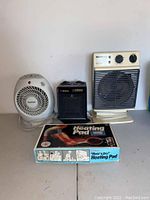 Photo of three electric heaters and boxed heating pad on a white surface against a white wall.