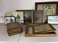 Wide view of framed artworks and antique books arranged on table, showing a religious portrait, landscape paintings, and art books.
