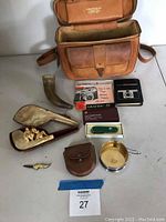 Overview photo showing all items including carved pipe with case, powder horn, Swiss Army knife, leather pouch, folding metal cup, camera manuals, and leather camera bag.