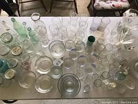 Top-down photo showing a large collection of assorted clear and tinted glass drinking glasses, mason jars, vintage soda bottles, plates and nesting bowls laid on two tables.