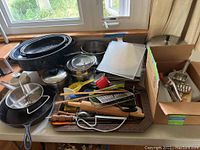 Full view of various kitchen pots, pans, utensils, cutting boards, and other kitchen accessories displayed on a table.