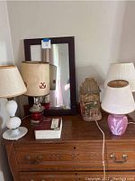 Five lamps in various styles and sizes arranged on a wooden dresser. One has a white hobnail milk glass base, another is a vintage-style lamp with anchor design on the shade, and includes a small pink ceramic lamp with leaf design. Also includes a small house-shaped book lamp and a wood framed rectangular mirror behind the lamps.