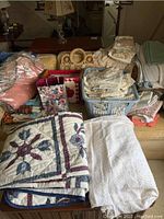 Lot includes quilted blankets with floral patterns, assorted linens including folded white and green fabric, lace and crocheted doilies, floral patterned cloths, placemats, and a set of wooden napkin rings.