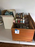 Side view of wooden box filled with textiles and cardboard box with fabric nearby, includes some thread spools on pegboard at rear
