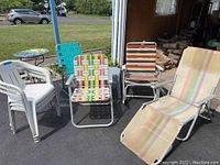 Outdoor set of various chairs and tables including plastic stacking chairs, two metal rocking chairs with colored webbing, fold-out lounge chair, plastic tables, and folding cot