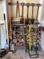 Wide view of the lot showing stacked firewood, short wooden ladder in front, green metal dolly, and various garden tools including long-handled shovels and rakes hung on a wooden board.