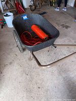 Black wheelbarrow with orange extension cord and roll of orange plastic sheeting inside, showing overall view of the items in the lot.
