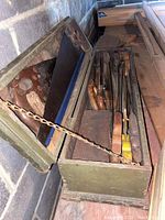 Open green wooden tool chest showing multiple hand saws with wooden handles inside, hinged lid held by a metal chain, placed against a concrete wall.