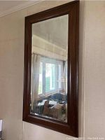 Rectangular wooden framed wall mirror reflecting room scene with window and furniture.