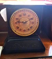 Front view of antique Ansonia mantel clock with cast iron body and gold gilt circular face showing clock hands and numerals.