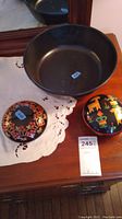 Photo showing the large black bowl and two smaller decorated lidded wood bowls placed on a wooden surface with a white cloth.
