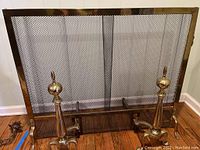 Full view of brass framed fireplace screen with black mesh and two brass andirons on hardwood floor.