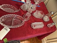 Multiple clear pressed glass serving pieces on red tablecloth, showing salt and pepper shakers, divided relish dish, butter saver, coasters, and other pieces