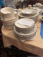 Stacked Lenox plates and bowls on a tan cloth-covered table as part of a dinnerware lot.