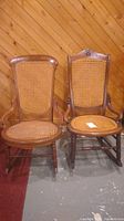 Side-by-side wooden rocking chairs with cane woven backs and seats. One chair with armrests, one armless. Chairs show carved details and scratches.