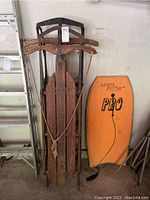 Two vintage wooden sleds stacked with rope tying them together, one sled marked Speedaway, alongside an orange Wave Action Pro boogie board.