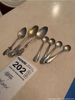 Seven small sterling silver spoons arranged side by side showing various detailed handles and tarnished surfaces, requiring cleaning.