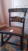 Full view of black wooden chair with cane seat and painted floral backrest, showing decorative details and overall condition.