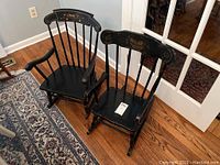 Two vintage child-sized black wooden rocking chairs with decorative paint on top rails shown side by side on hardwood floor near doorway and rug.