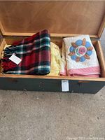Photo showing open trunk containing folded red plaid blanket, yellow quilt, and floral applique quilt all vintage linens.