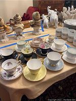 Photo showing 11 assorted vintage tea cups and saucers arranged on a table with decorative elephants and figurines in the background.