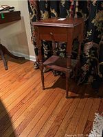 Full view of the vintage wood side table showing turned legs, drawer with knob, and lower shelf on hardwood floor with patterned curtain background.