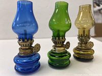 Three mini oil lamps in cobalt blue, green, and amber colors with glass bases and chimneys featuring brass wick adjusters.