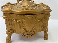 Front angle view of an ornate gold gilt jewelry box showing detailed relief decorations and curled legs.