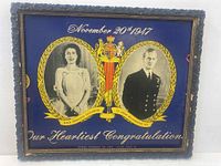 Full view of carved wooden framed picture with central colored coat of arms and oval portraits of Princess Elizabeth and Prince Philip on blue background.