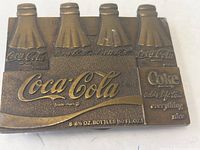 Close up of brass belt buckle showing embossed Coca Cola bottles and logo