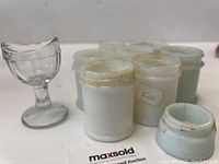 Photo showing the clear glass eye wash cup alongside six small white milk glass jars with lids, some jars have mild staining.