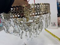 Close-up of gold-tone metal circular frame with teardrop crystal drops hanging from it, showing size comparison with a hand and tape measure.