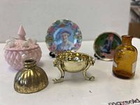 Miniature dollhouse accessories including pink glass jar with white bird accents, brass bowl with ornate legs, amber bottle, and two decorative miniature plates