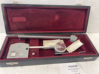 Planimeter and accessories stored in a fitted black textured case with red velvet lining, showing the entire device, magnifying glass, accessory, and papers.