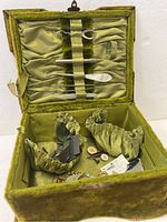 Open green plush Victorian sewing box showing satin-lined interior with sewn-in elastic bands holding metal and mother-of-pearl sewing tools, fabric pincushions, buttons, and other sewing accessories inside.