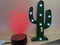 Photo showing both the wolf face LED light (red) and the cactus marquee light with white bulbs on a surface against a wall.
