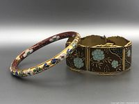 Photo showing two cloisonné bracelets, one round floral bangle and one wider hinged bracelet with blue flowers on brown background.