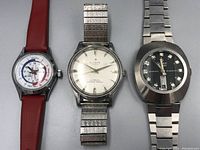 Front view of three mechanical wristwatches side by side: Time Trainer on left with red band, Alecya in center with expansion band, and Omax on right with black dial and metal bracelet.