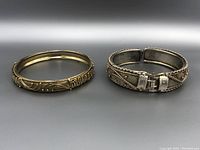 Both vintage silver bracelets shown side by side on a white-gray surface. Left bracelet is a slip-on bangle with ornate designs. Right bracelet is hinged with a clasp.