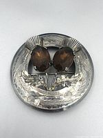 Front view of round silver Scottish kilt pin with two oval faceted citrine gemstones set on detailed thistle stems with engraved thistles around the edge.