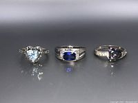 Front view of three sterling silver rings with different blue and multicolor heart and rectangular gemstones on a solid surface.