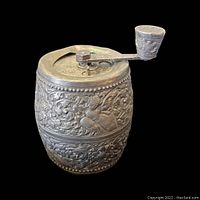 Front angled view showing the hand crank and embossed floral and figure design on the body of the peppermill.