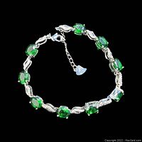 Sterling silver tennis bracelet displaying nine oval green gemstones and clear gemstone links, laid flat against a black background.