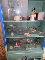 Glass storage jar, ceramic pitcher, various glass bowls, a porcelain doll, metal candle holder and decorative items on teal shelves.
