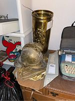 Photo showing the brass umbrella stand, metal helmet with decorative trim placed on a wooden surface with other miscellaneous items nearby.