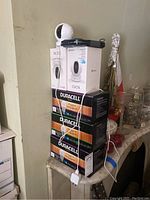 Stack of Ezviz C6CN camera boxed, waterproof phone case, and three boxes of Duracell Ultra light bulbs on table.