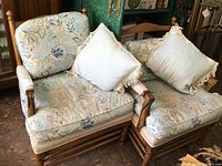 Two matching upholstered chairs with wooden frames. Light blue floral and paisley pattern cushions and backs. Each has a matching light blue pillow with fringed edges. Chairs show sun fading and wear.